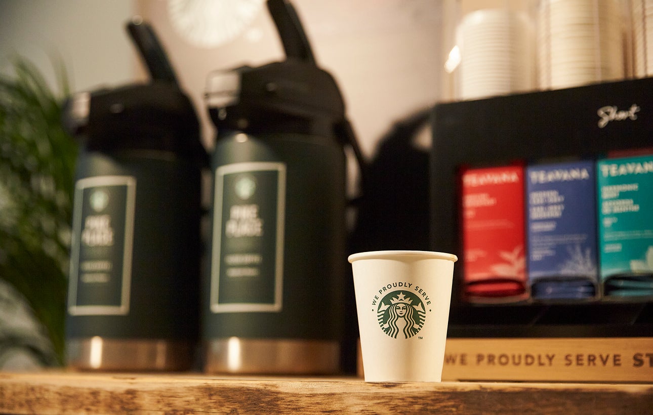 Et We Proudly Serve Starbucks-papbæger står på en trædisk foran store automater med færdigbrygget kaffe