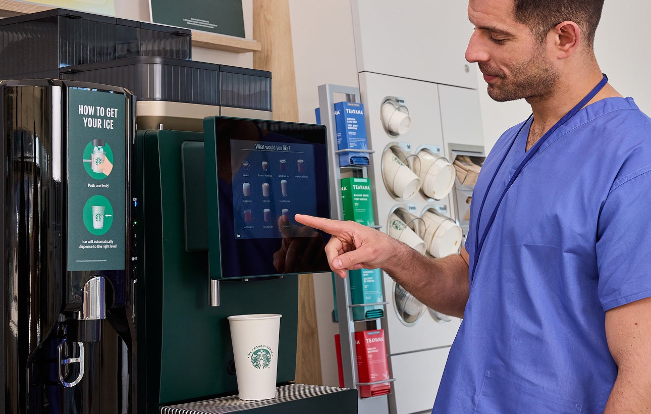 Sundhedspersonale vælger en drik ved en We Proudly Serve Starbucks-kaffeautomat med selvbetjening. 
