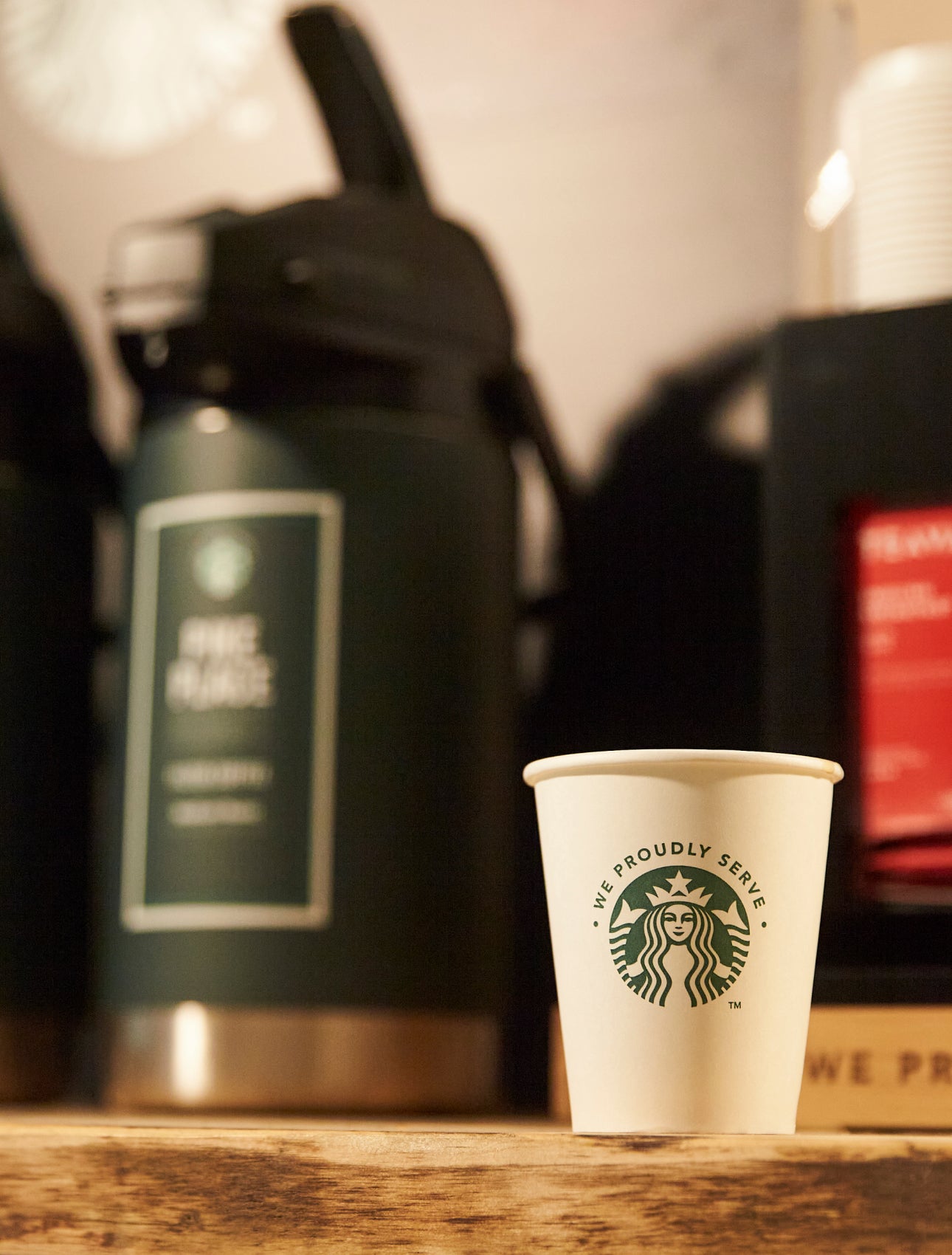 Et We Proudly Serve Starbucks-papbæger står på en trædisk foran store automater med færdigbrygget kaffe