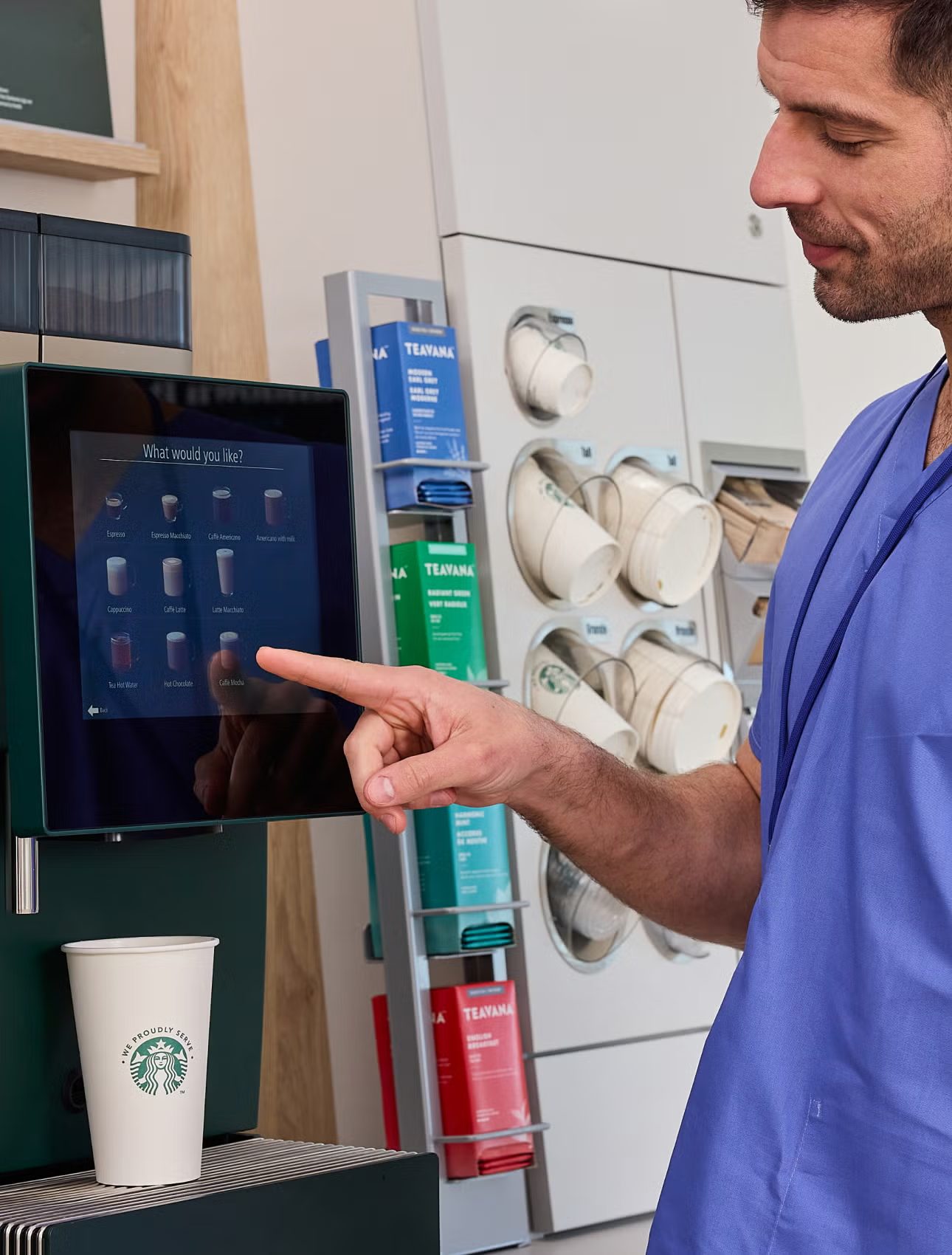 Sundhedspersonale vælger en drik ved en We Proudly Serve Starbucks-kaffeautomat med selvbetjening. 