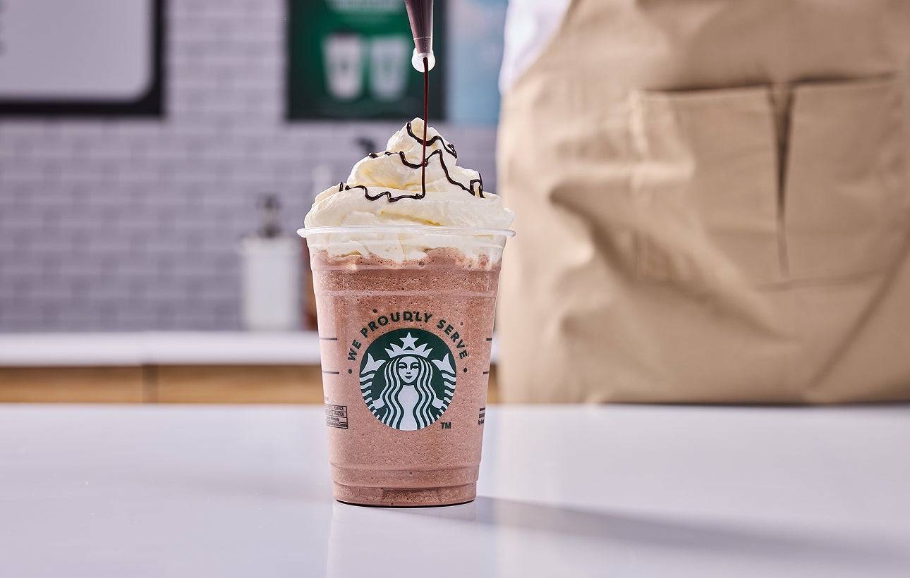 En We Proudly Serve Starbucks-drik toppet med flødeskum og en barista, der drysser chokolade ovenpå.