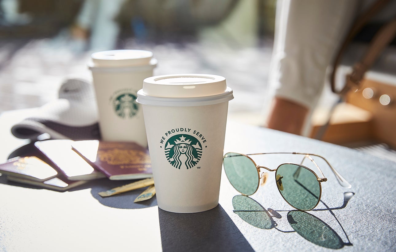 To We Proudly Serve Starbucks-takeawaybægre med låg placeret på et udendørs bord ved siden af et par solbriller