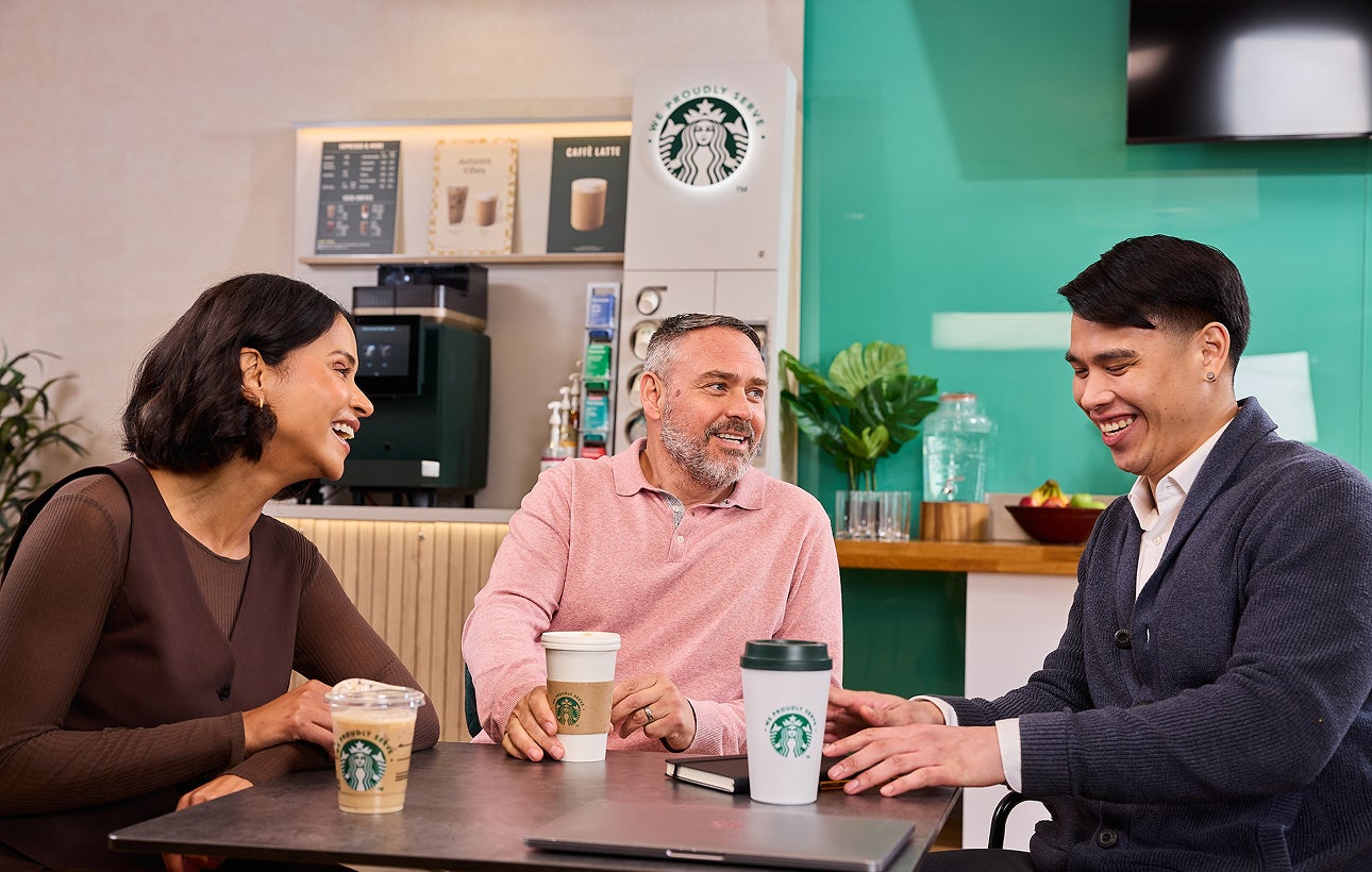 Kolleger sidder sammen med We Proudly Serve Starbucks-bægre og en kaffeautomat med selvbetjening i baggrunden.