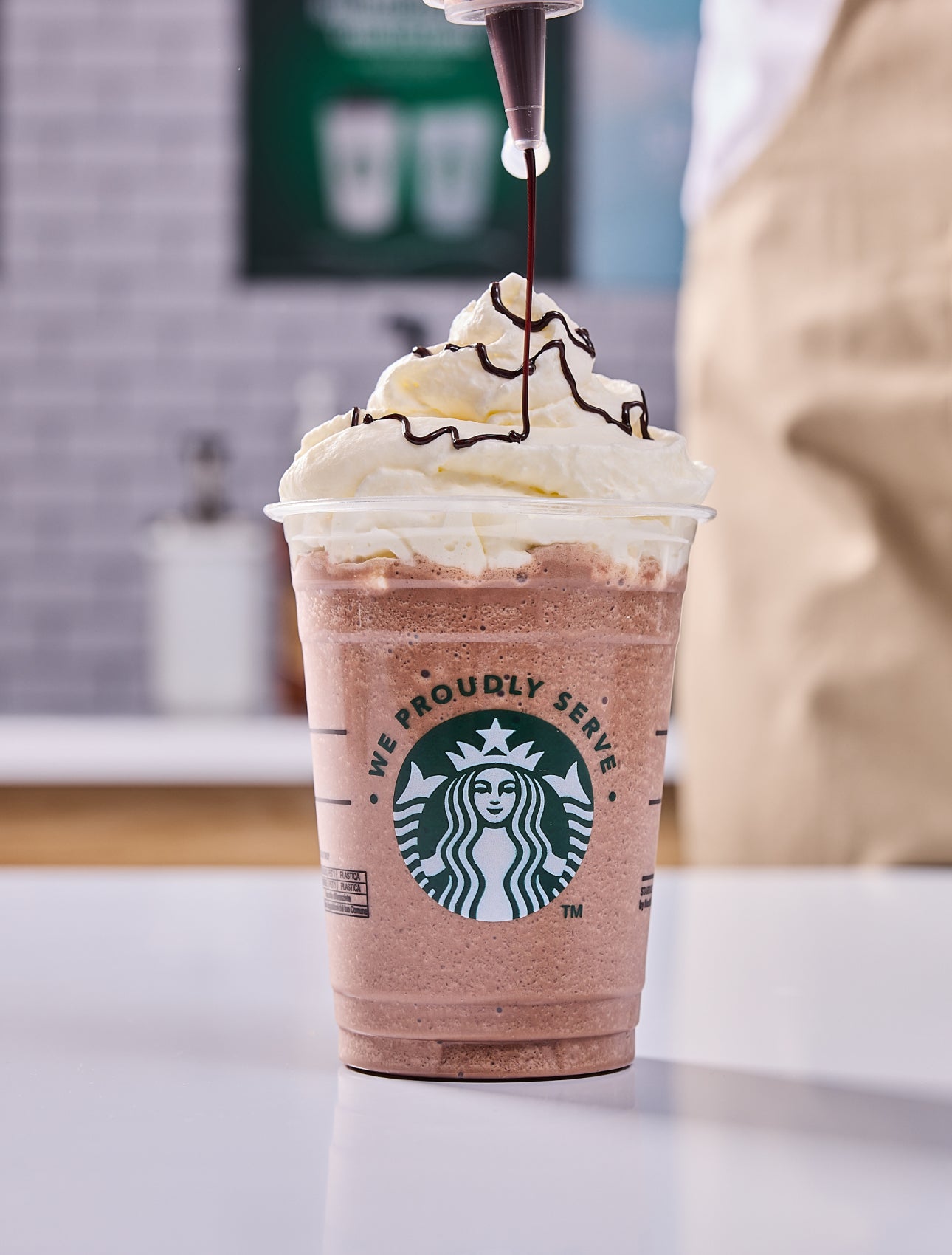 En We Proudly Serve Starbucks-drik toppet med flødeskum og en barista, der drysser chokolade ovenpå.