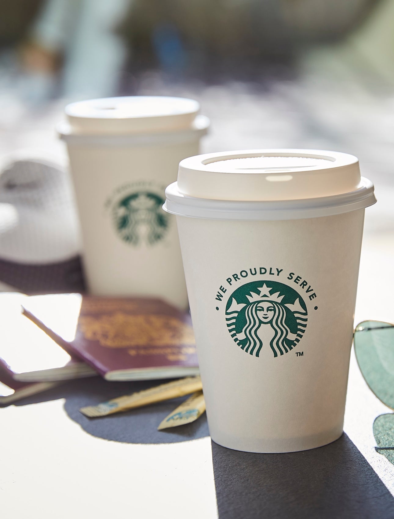 To We Proudly Serve Starbucks-takeawaybægre med låg placeret på et udendørs bord ved siden af et par solbriller