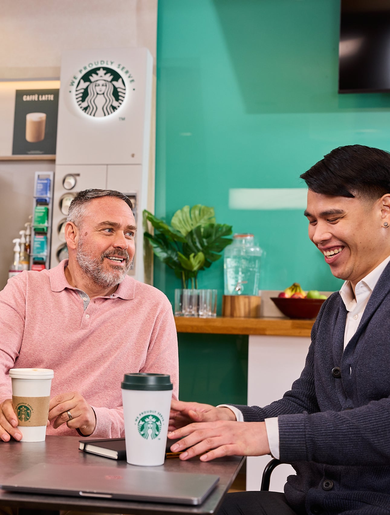 Kolleger sidder sammen med We Proudly Serve Starbucks-bægre og en kaffeautomat med selvbetjening i baggrunden.