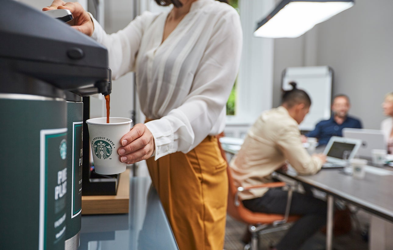 En person fylder et We Proudly Serve Starbucks-papbæger med færdigbrygget kaffe fra en kaffeautomat