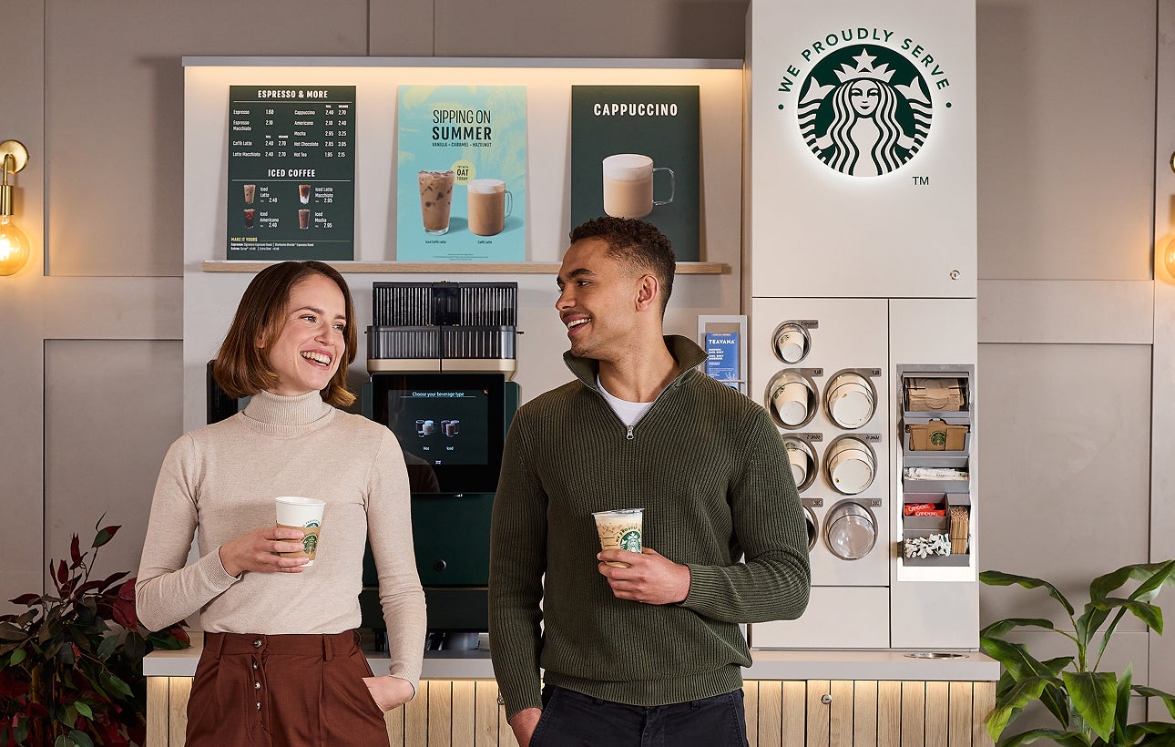 To personer står og snakker, mens de holder We Proudly Serve Starbucks-bægre foran en kaffestation med selvbetjening. 