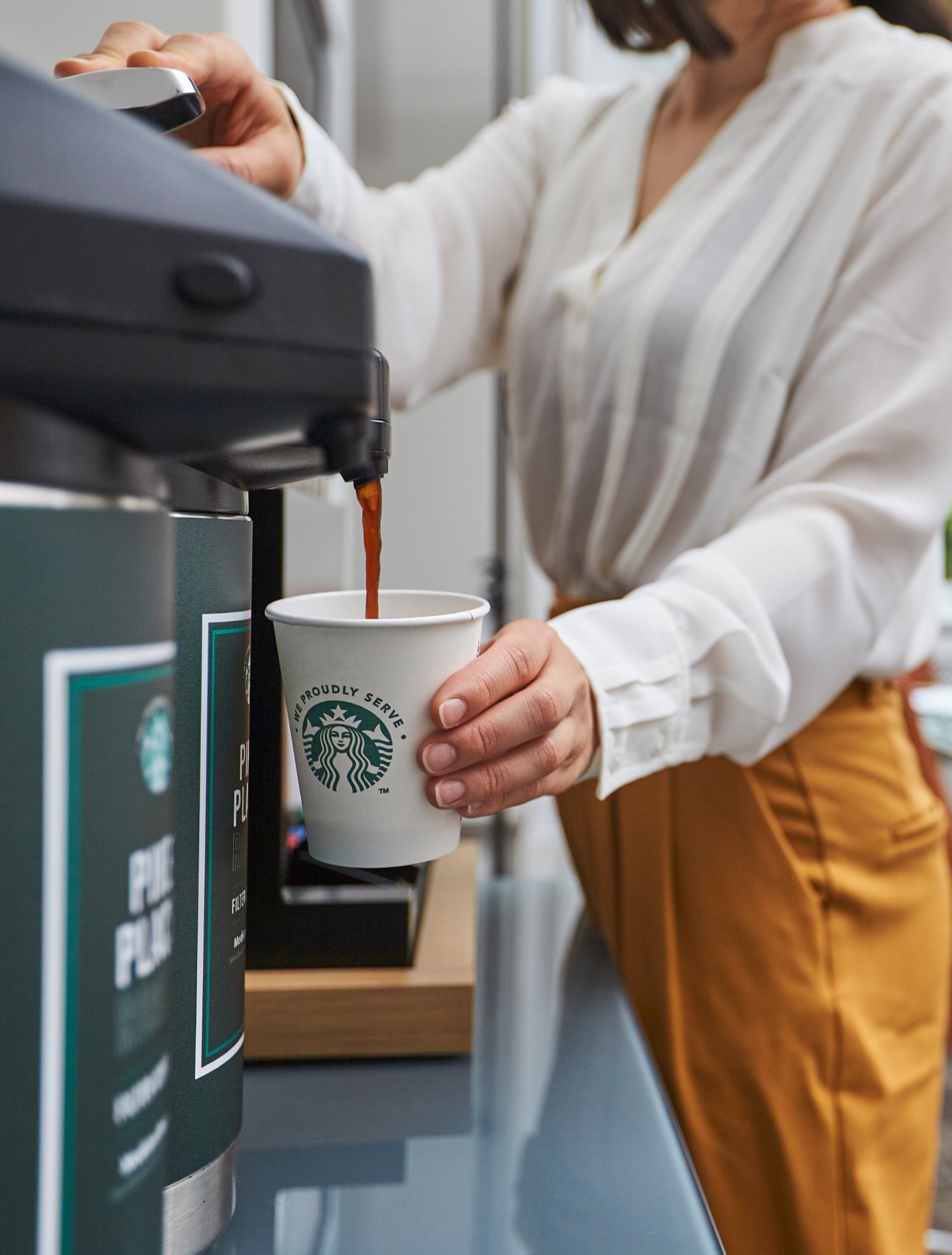 En person fylder et We Proudly Serve Starbucks-papbæger med færdigbrygget kaffe fra en kaffeautomat