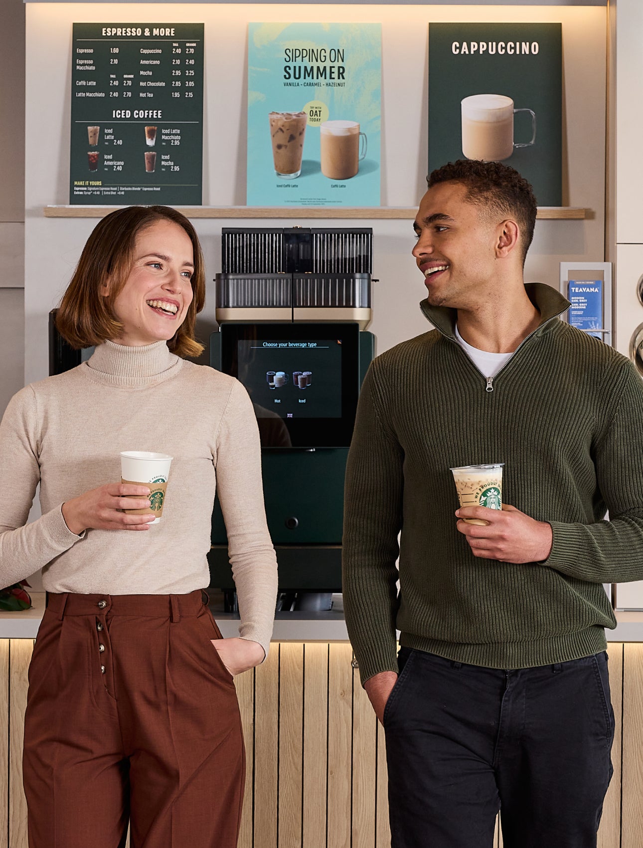 To personer står og snakker, mens de holder We Proudly Serve Starbucks-bægre foran en kaffestation med selvbetjening. 