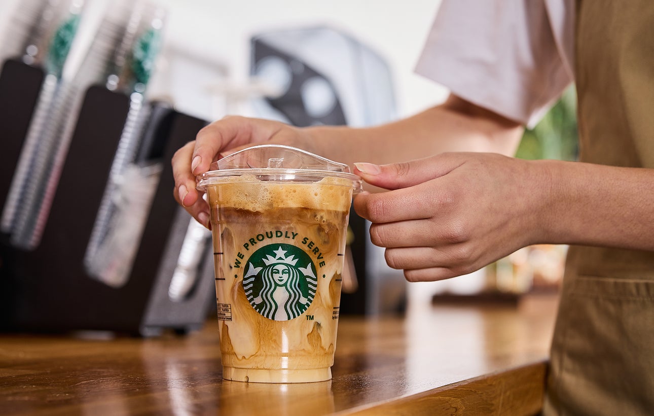 En barista sætter låg på en We Proudly Serve Starbucks-iskaffe med hvirvler af fløde og kaffe