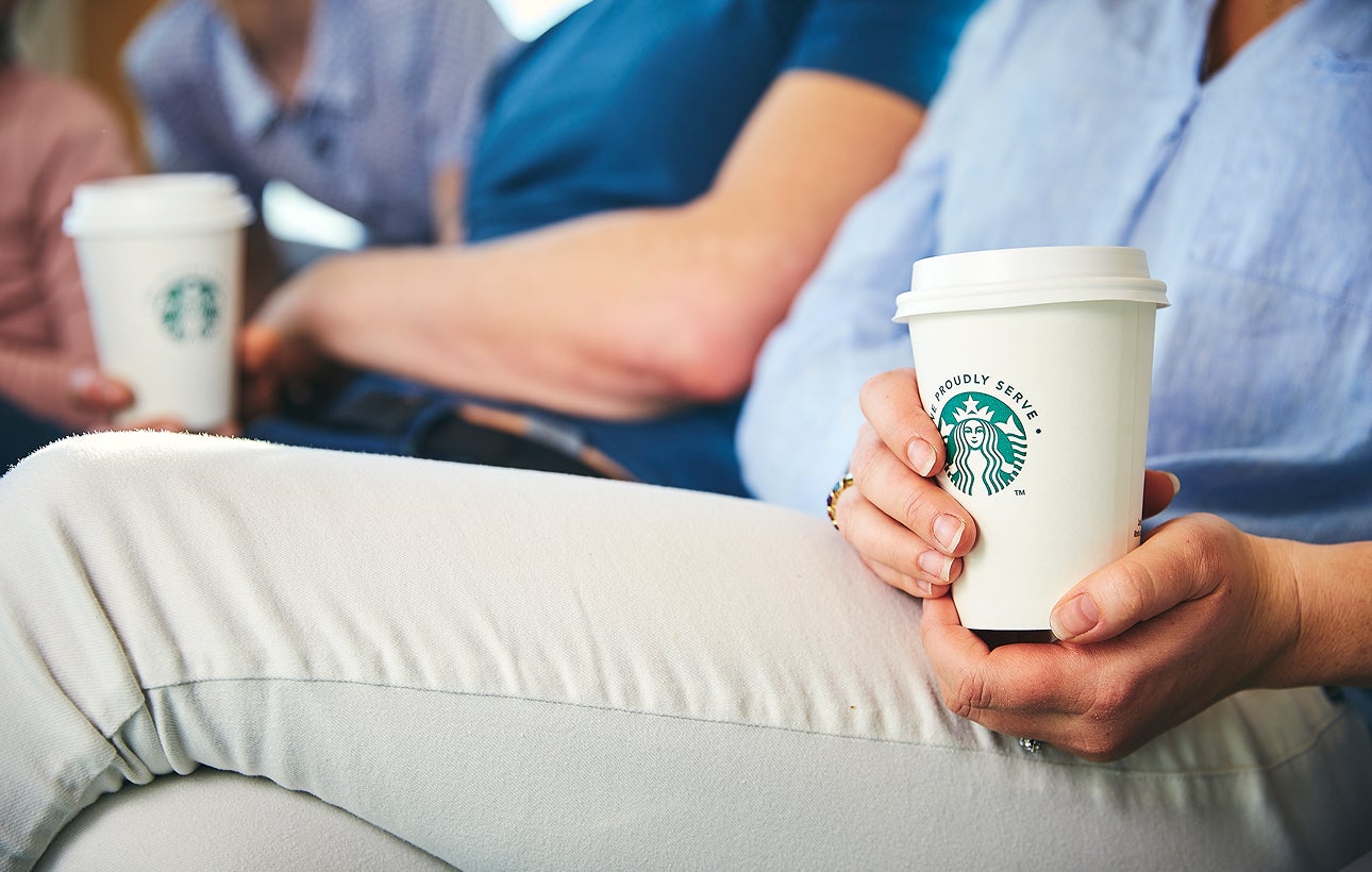 En person sidder ned og holder et We Proudly Serve Starbucks-takeawaybæger med begge hænder