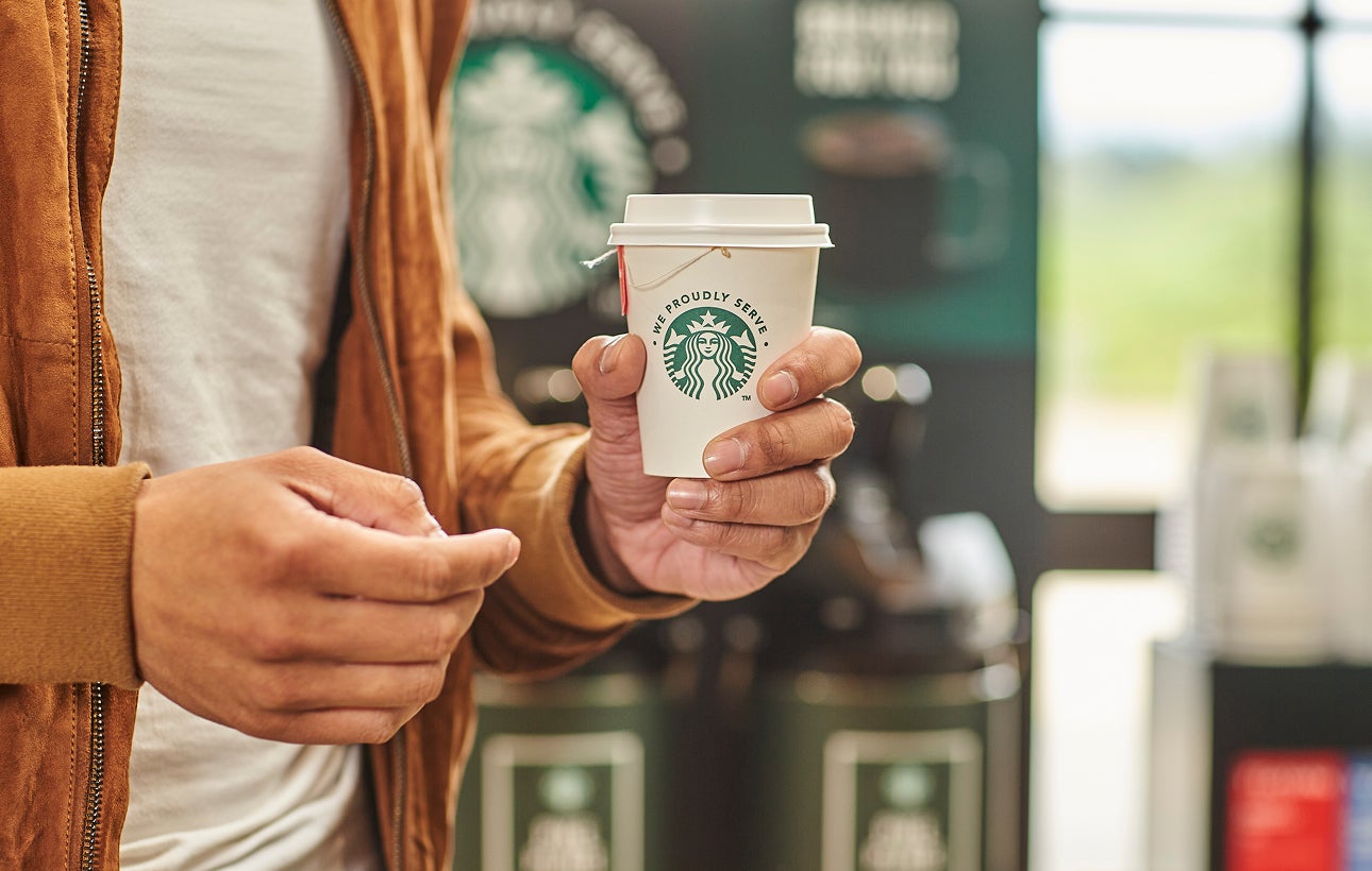 En person holder en kop te i et We Proudly Serve Starbucks-takeawaybæger