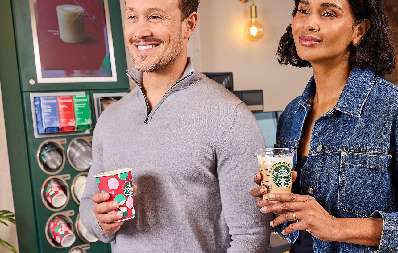 En mand, der holder en We Proudly Serve Starbucks-kaffekop med et logo, der passer til højtiden, og en kvinde, der holder en 