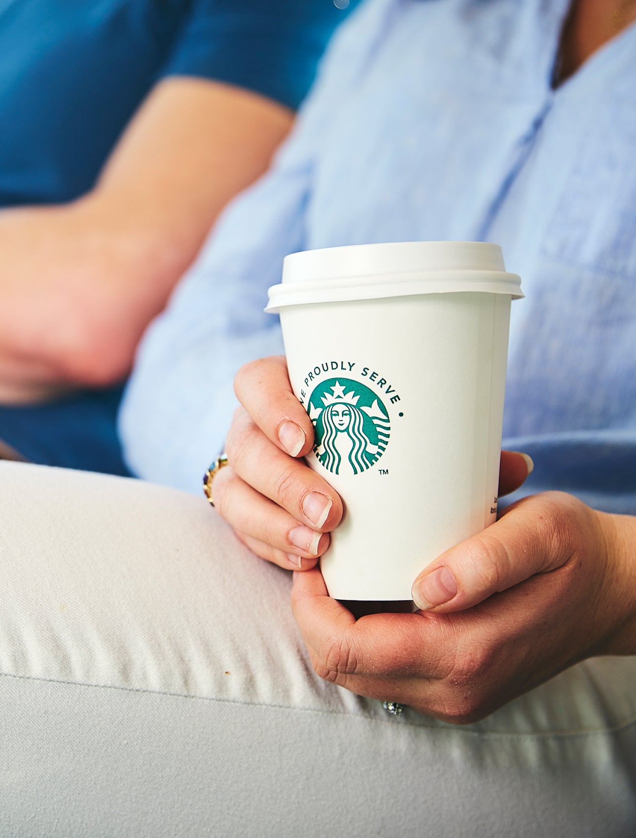 En person sidder ned og holder et We Proudly Serve Starbucks-takeawaybæger med begge hænder