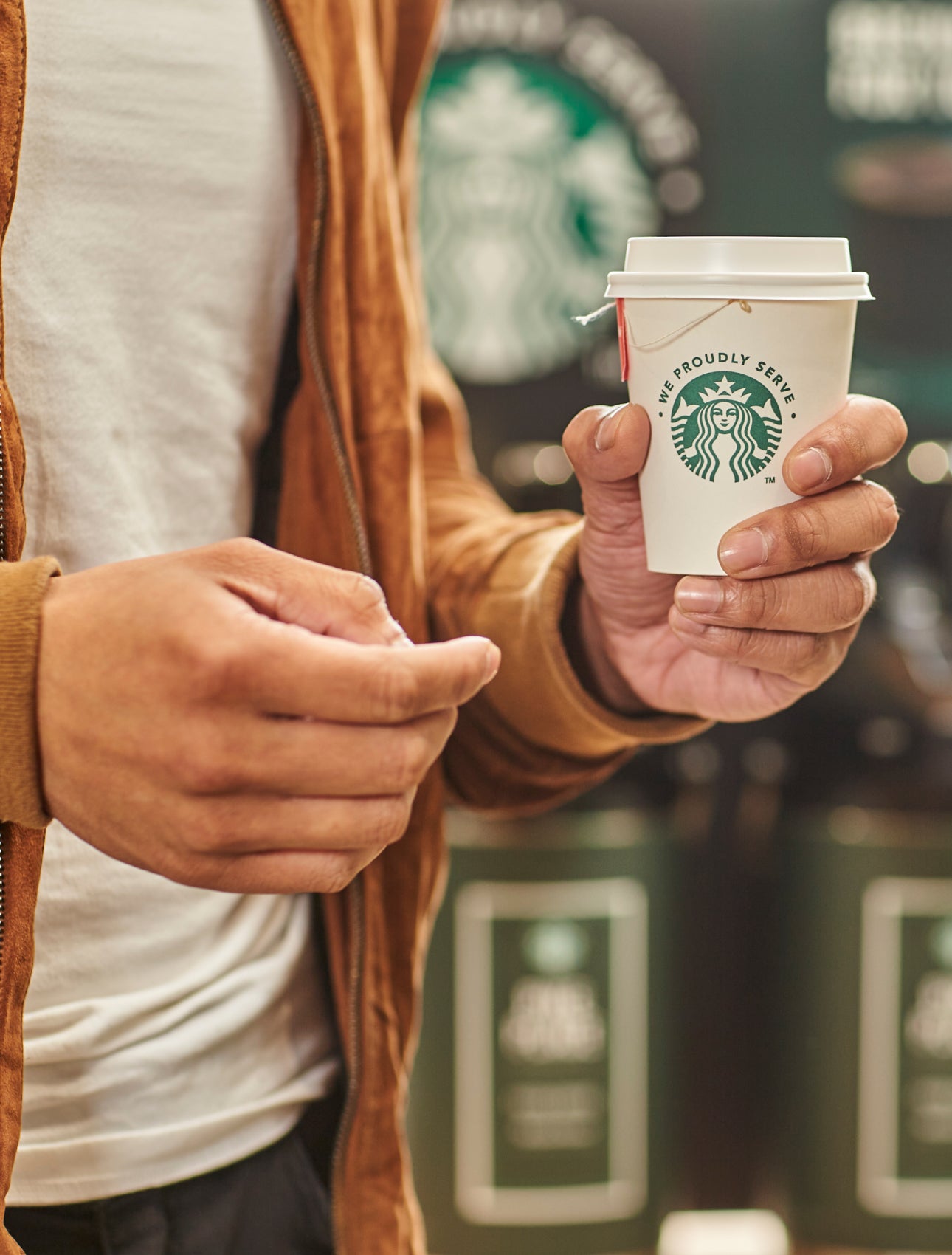 En person holder en kop te i et We Proudly Serve Starbucks-takeawaybæger