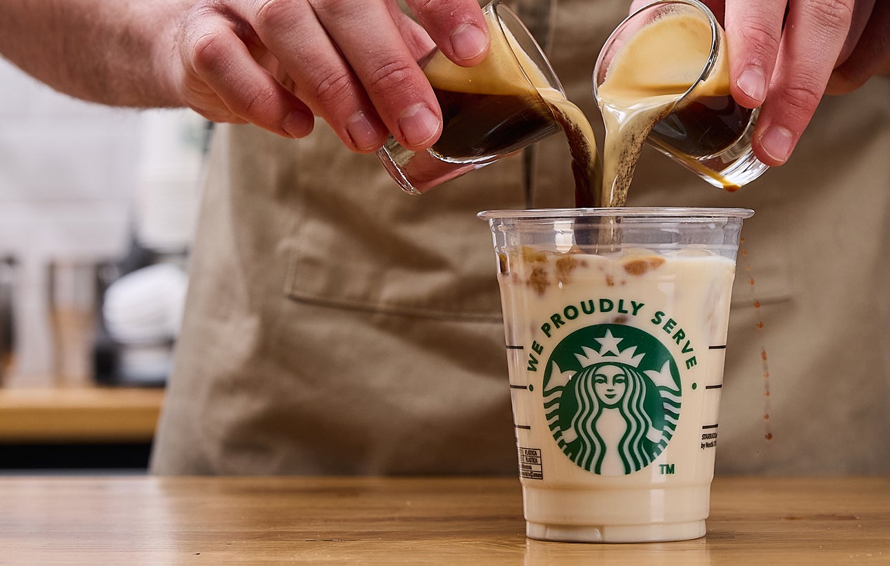 En barista skænker to espressoshots samtidig i et We Proudly Serve Starbucks-bæger fyldt med is og mælk