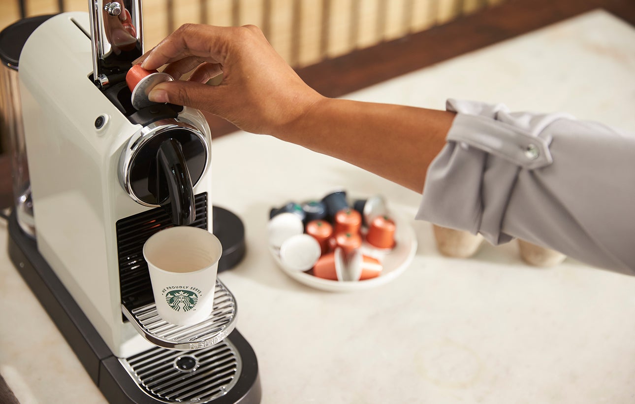 En person sætter en kaffekapsel i en kaffemaskine til enkeltservering med et We Proudly Serve Starbucks-bæger på drypbakken.