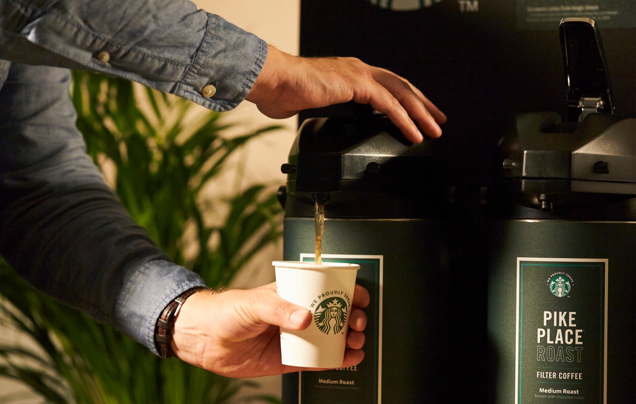 En person serverer Starbucks Pike Place® Roast-kaffe fra en stor kaffeautomat i et We Proudly Serve Starbucks-bæger