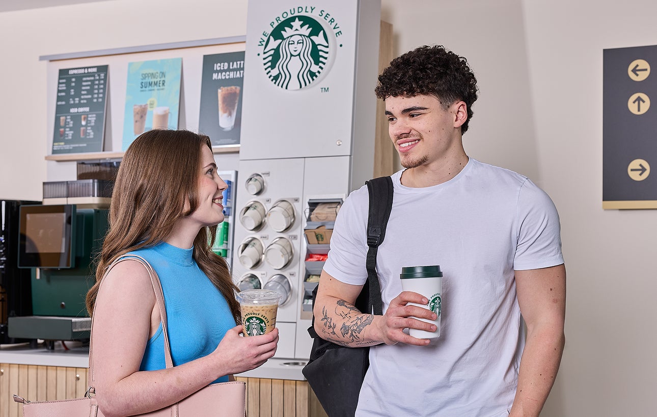  En kvinde og en mand står foran en We Proudly Serve Starbucks-kaffestation med selvbetjening