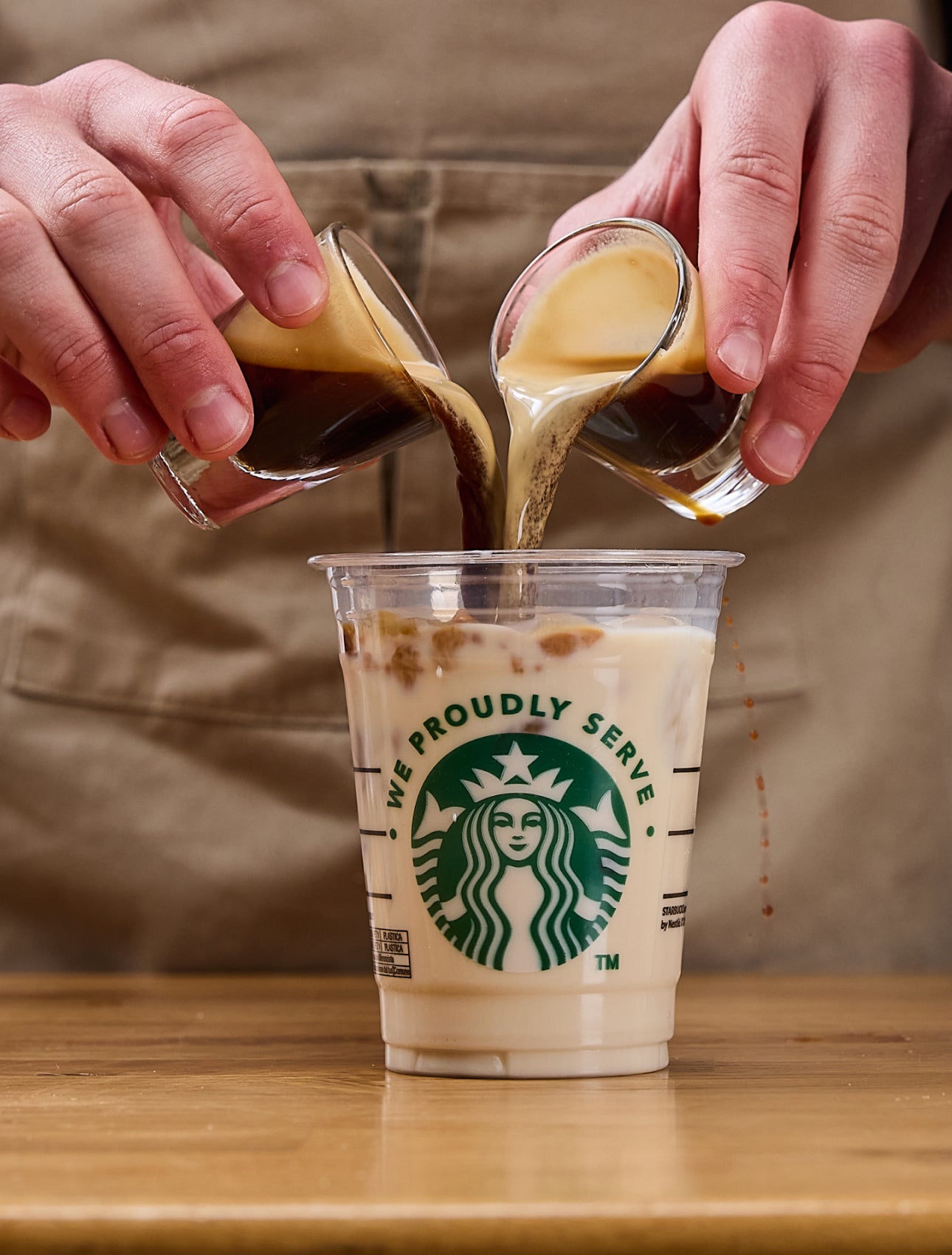 En barista skænker to espressoshots samtidig i et We Proudly Serve Starbucks-bæger fyldt med is og mælk