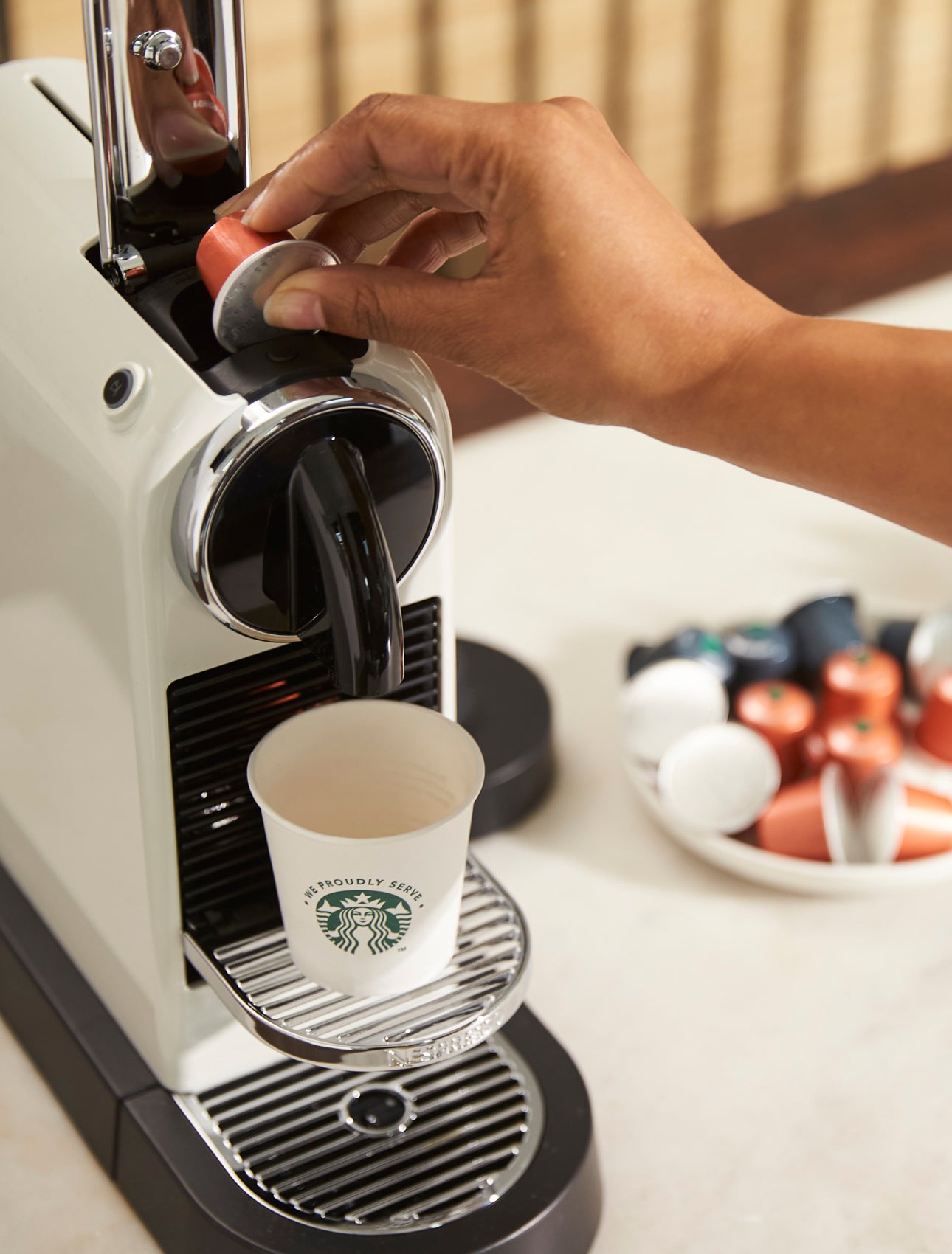 En person sætter en kaffekapsel i en kaffemaskine til enkeltservering med et We Proudly Serve Starbucks-bæger på drypbakken.