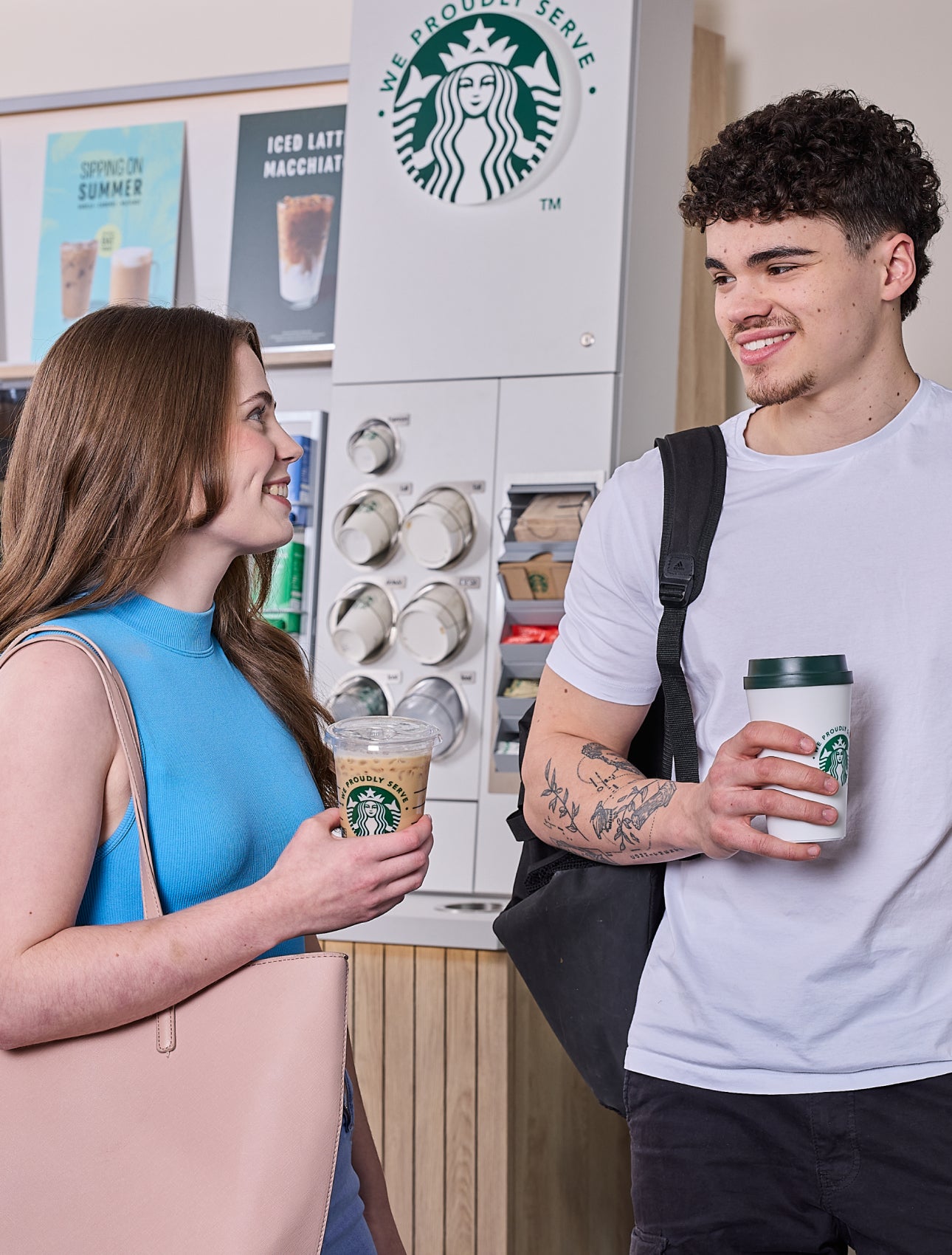  En kvinde og en mand står foran en We Proudly Serve Starbucks-kaffestation med selvbetjening