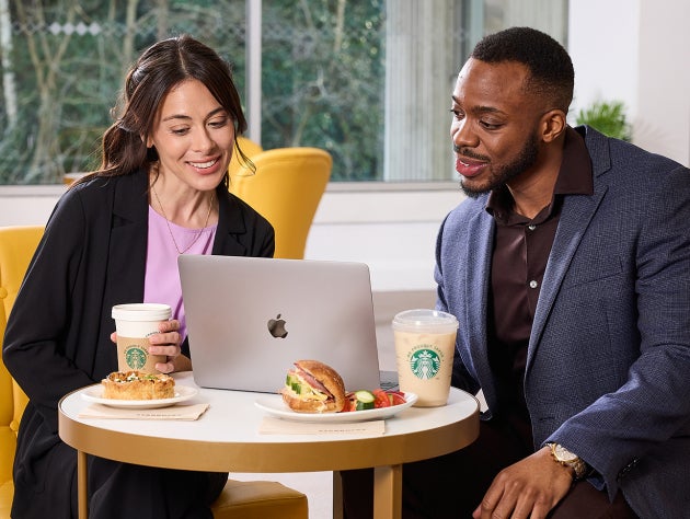 To personer sidder foran en bærbar computer med We Proudly Serve Starbucks-kaffe og sandwich på bordet.