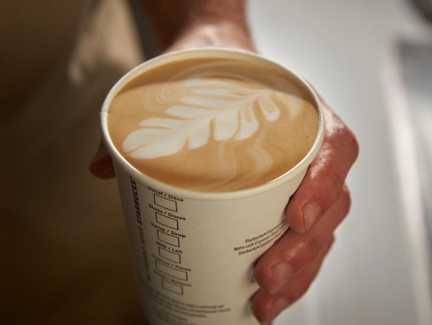 En hånd, der holder en kop We Proudly Serve Starbucks-kaffe med latte art på toppen. 
