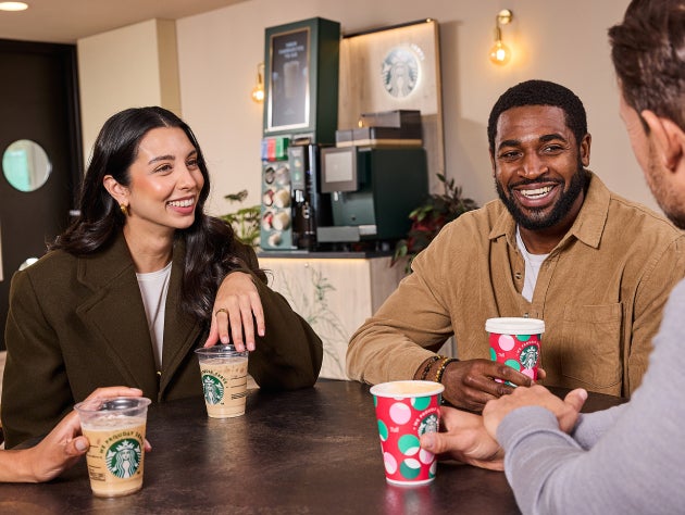 Fire personer sidder smilende ved et cafébord med Starbucks-drikke i festlige og standardiserede We Proudly Serve Starbucks-b
