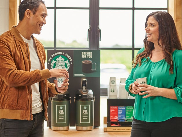 To personer står og snakker, mens de holder We Proudly Serve Starbucks-bægre foran automater til færdigbrygget kaffe
