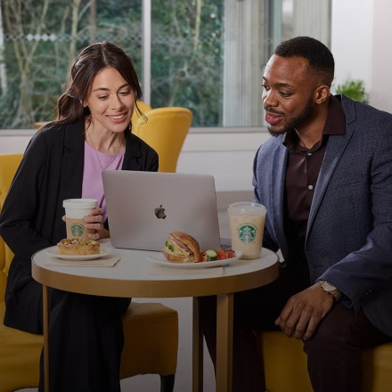 To personer sidder foran en bærbar computer med We Proudly Serve Starbucks-kaffe og sandwich på bordet.