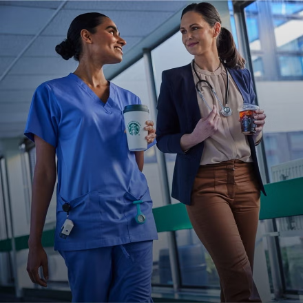 To sundhedsmedarbejdere går sammen på et hospital. De smiler og har "We Proudly Serve Starbucks"-drikke i hånden. 
