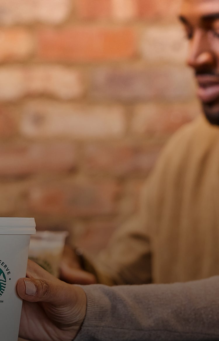 En person, der holder en We Proudly Serve Starbucks-kop foran en sløret murstensbaggrund 