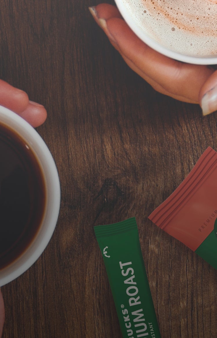 To hænder holder en sort kaffe og en skummende cappuccino på et bord ved siden af Starbucks® Premium Instant-kaffebreve 