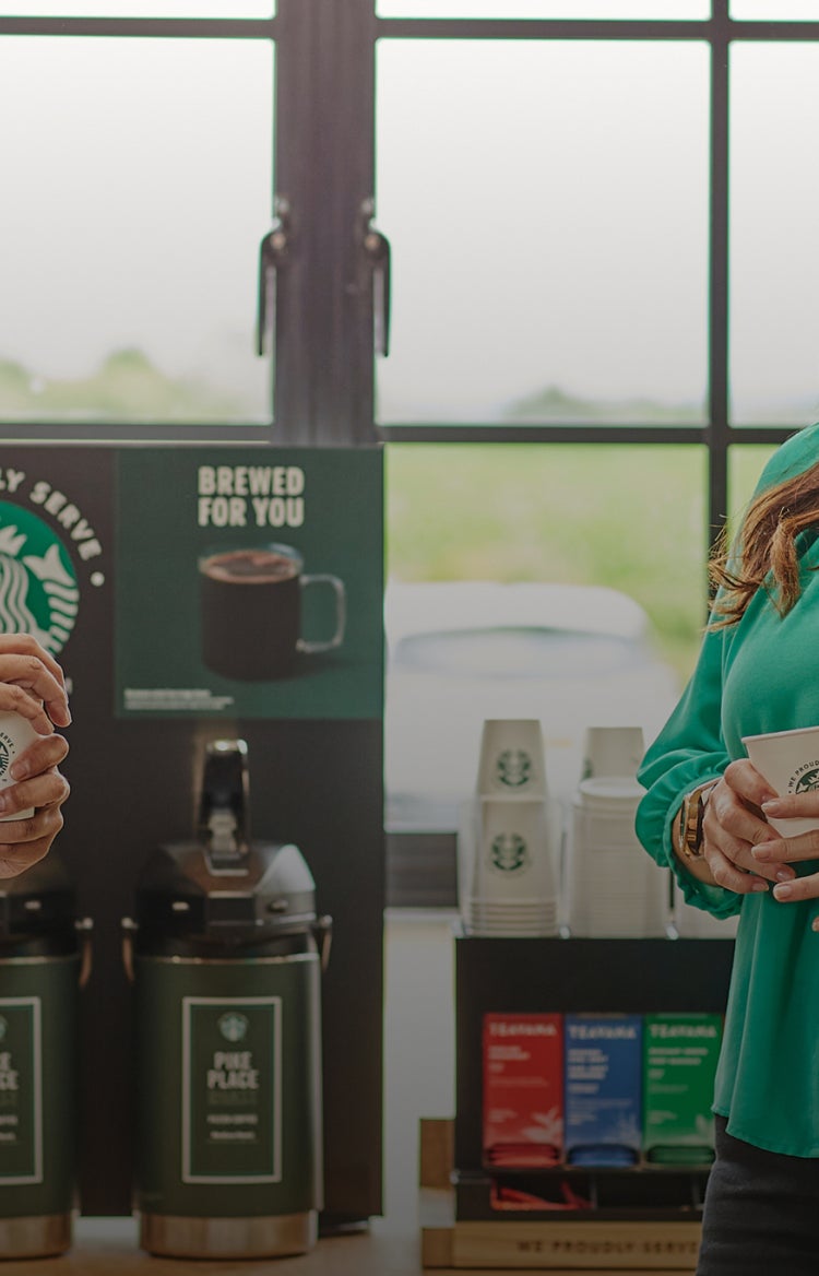 To personer står og snakker, mens de holder We Proudly Serve Starbucks-bægre foran automater til færdigbrygget kaffe