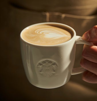 En hånd, der holder et hvidt Starbucks-krus fyldt med en latte, toppet med hjerteformet latte art