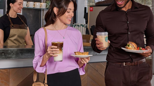 En kvinde og en mand står ved en cafédisk på arbejdspladsen og smiler, mens de henter iskaffe og mad fra Starbucks