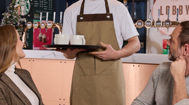 En barista serverer to Starbucks-drikke til kunder i en hotellobbybar med en We Proudly Serve Starbucks®-menu bagved.