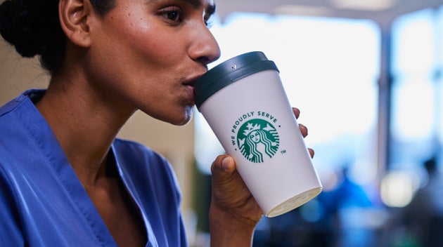 En sundhedsmedarbejder i blå kittel drikker en kop We Proudly Serve Starbucks-kaffe, mens vedkommende sidder i en personaleka