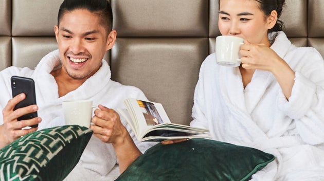 Et par slapper af på en hotelseng i hvide morgenkåber, mens de nyder en kop Starbucks-kaffe.  