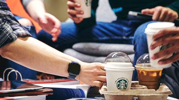 En gruppe studerende omkring et bord med bøger og notesbøger, hvor en af de studerende tager en We Proudly Serve Starbucks-ka