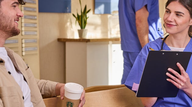En kvinde i en blå kittel taler med en patient, der holder en We Proudly Serve Starbucks-kaffekop i hænderne, på en klinik. 
